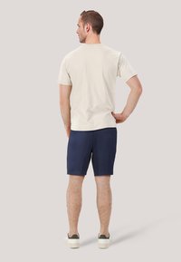 Beige bomullströja med rund halsringning, tillsammans med marinblå shorts och avslappnade vita sneakers med gröna detaljer. Utsikt från baksidan.