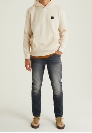 Sweat à capuche crème en tissu texturé, avec poche kangourou et écusson logo, porté sur une chemise moutarde. Un jean bleu foncé et des baskets beiges complètent la tenue.