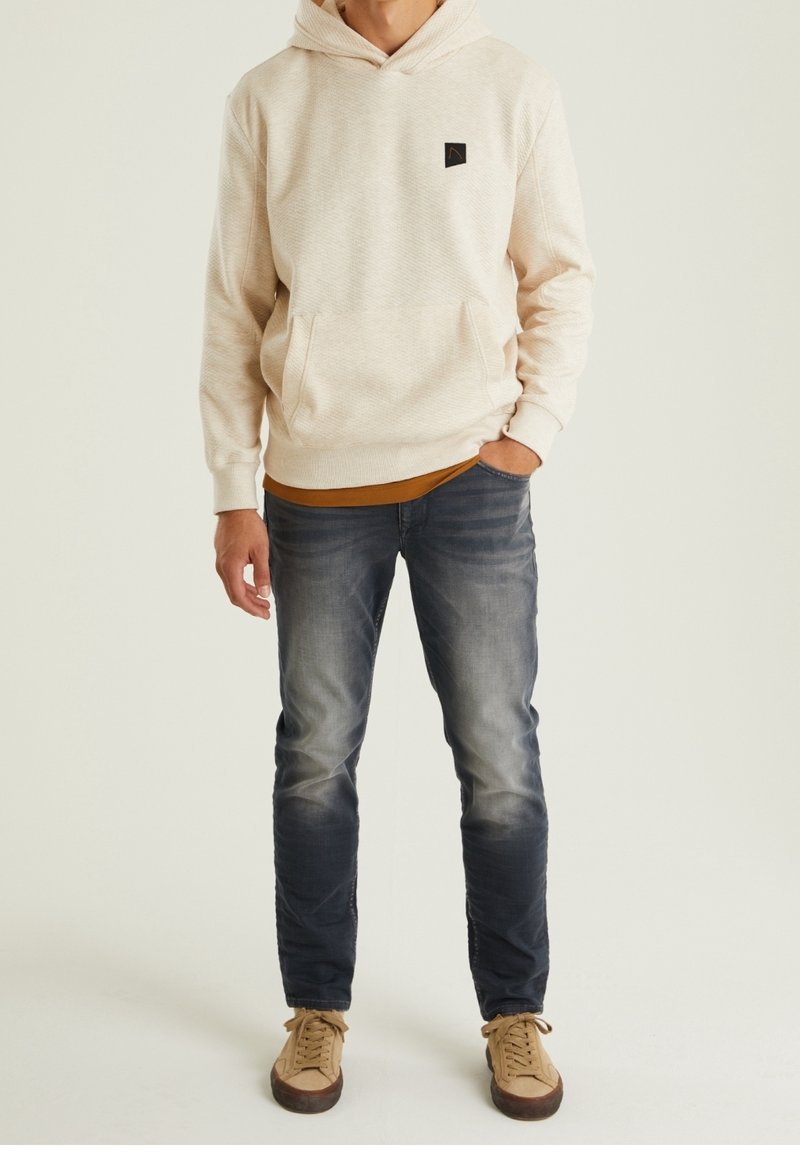 Sweat à capuche crème en tissu texturé, avec poche kangourou et écusson logo, porté sur une chemise moutarde. Un jean bleu foncé et des baskets beiges complètent la tenue.