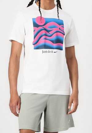 Person iført hvid t-shirt med pink og blå abstrakt design og teksten "Just do it", kombineret med lysegrå shorts med lille logo.