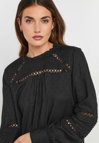 Blusa nera in tessuto texturizzato, con dettagli in pintuck e pizzo sulle spalle e sulle maniche, e una finitura arricciata al collo.