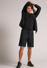 Czarna kurtka na zamek błyskawiczny noszona na czarnym T-shircie, w zestawieniu z czarnymi szortami z niebieskimi akcentami i strukturalnym wzorem. Uzupełnione czarnymi sneakersami.