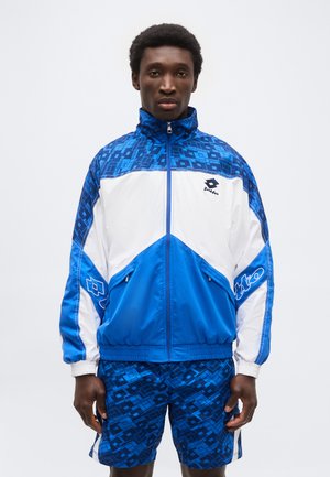 Meesmodell kannab sinise ja valge geomeetrilise mustriga jopet ning sobivaid siniseid mustrilisi lühikesi pükse ühevärvilise tausta ees.