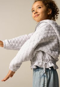 Veste grise matelassée avec capuche, présentant des poignets côtelés et un ourlet à cordon de serrage ajustable, accompagnée d'un jean taille haute bleu clair.