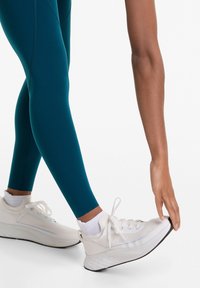 Törökzöld leggings könnyű, fehér, hálós paneles és fűzős sportcipővel; rugalmas talp hangsúlyozza a mozgékonyságot, miközben nyújt.