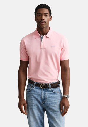 Man wearing a light pink polo shirt tucked into light blue jeans with a dark braided belt and silver wristwatch.