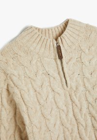 Suéter de punto trenzado color crema con un alto cuello acanalado y cremallera de cuarto, que presenta una pequeña etiqueta de cuero en la cremallera y una textura suave.