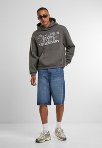 Sudadera gris con capucha y texto blanco, pantalones cortos de mezclilla, calcetines blancos y zapatillas grises. El modelo lleva gafas de sol y se presenta con confianza.