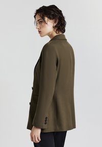 Frau mit kurzem nassen dunklem Haar, die ein olivgrünes Blazer und schwarze Hosen trägt, im Profil vor einem schlichten hellen Hintergrund.