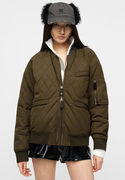 Νεαρή γυναίκα που φοράει μια επενδεδυμένη μπουφάν ελιάς, μαύρα βινύλια σορτς και γκρι καπέλο με μαύρες γούινες στα αυτιά, σε λευκό φόντο.