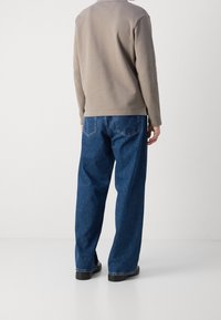 Person wearing loose blue jeans, taupe long-sleeve shirt, and black shoes standing against a white background, back facing camera.