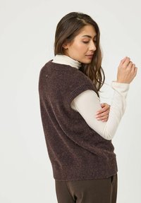 Pull sans manches en tricot marron avec une finition texturée, ourlet côtelé et col arrondi, porté par-dessus un haut à manches longues de couleur claire.