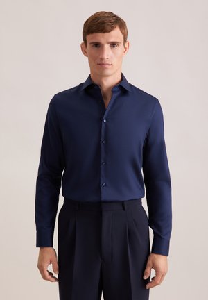 Marineblaue Langarmbluse aus glattem Stoff, mit klassischem Kragen und durchgehender Knopfleiste, kombiniert mit dunklen, gefalteten Hosen.
