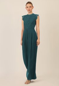 Teal jumpsuit met een kanten top met scalloped randen en korte mouwen, vloeiende broeken en zijplooien. Transparante hakken maken de look compleet.
