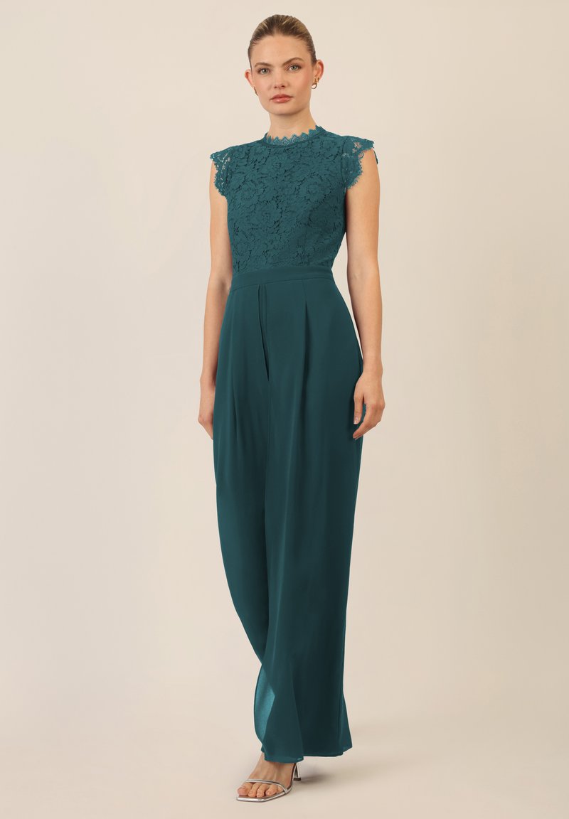 Teal jumpsuit met een kanten top met scalloped randen en korte mouwen, vloeiende broeken en zijplooien. Transparante hakken maken de look compleet.