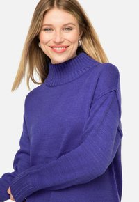 Lila gestrickter Pullover mit hohem Rippkragen, lockerer Passform und langen Ärmeln. Strukturiertes Muster verstärkt die visuelle Anziehungskraft. Keine sichtbaren Verzierungen.