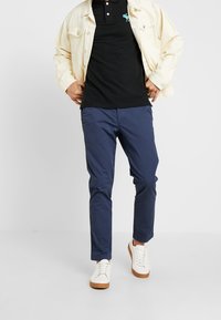 Chino blu navy con vestibilità slim, abbinati a una polo nera e a una giacca di denim crema. Calzature: sneakers bianche con dettagli color nocciola.