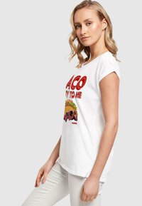 Hvid kortærmet t-shirt med rød grafisk tekst og billeder af en taco og en karakter, samt en glat, let tekstur.