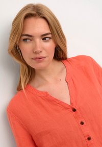 Koralinė lino marškinė su V formos iškirpte, turinti tris rudus mygtukus ir trumpas rankoves. Audinys turi švelnų, tekstūruotą išvaizdą.