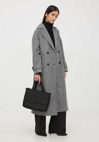 Cappotto grigio doppiopetto con bottoni grandi, indossato su un dolcevita nero. La modella tiene in mano una borsa a tracolla imbottita nera e indossa pantaloni neri a gamba larga.