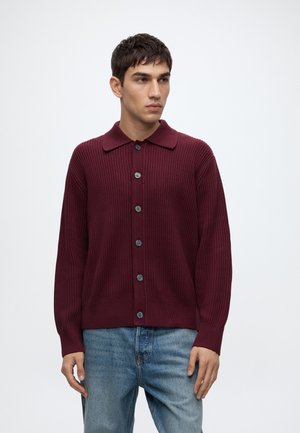 Jeune homme portant un cardigan en maille bordeaux boutonné et un jean bleu, debout devant un fond blanc uni, regardant vers sa gauche.