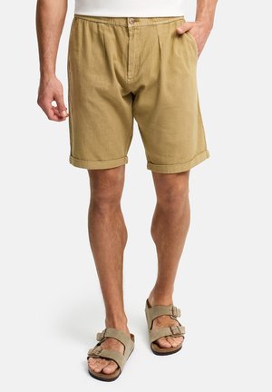 Mann trägt hellbraune Freizeitshorts mit aufgekrempelten Säumen und beigefarbene Sandalen mit Schnallen, steht mit einer Hand in der Tasche.