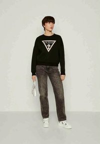 Μαύρη φούτερ μπλούζα με τριγωνικό λογότυπο που περιλαμβάνει το κείμενο "GUESS"· συνδυασμένη με σκούρα γκρίζα τζην παντελόνι και λευκά αθλητικά παπούτσια. Το μοντέλο κρατά μια μικρή τσάντα.