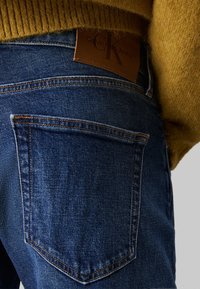 Jeans din denim de un albastru închis, cu o etichetă din piele Calvin Klein, având buzunare spate clasice și accente de cusătură vizibile.
