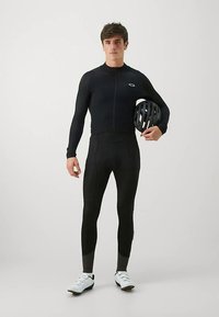 Svart cykeloverall med långa ärmar och dragkedja fram, kombinerad med svarta cykelbyxor. Modellen håller i en svart hjälm och bär vita cykelskor.