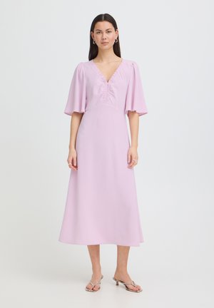 Frau trägt ein hellviolettes Midikleid mit flatternden Ärmeln, V-Ausschnitt und Rüschendetail, kombiniert mit offenen Sandalen mit niedrigem Absatz.