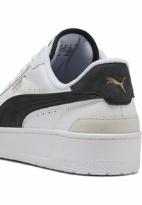 White sneaker with black accents, textured leather panel, gold logo, and smooth rubber sole. Rear features a suede-like patch and branding.
