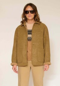 Veste en suédine marron avec fermeture boutonnée, deux poches avant, et un pull texturé beige en dessous. Portée avec des lunettes de soleil foncées.