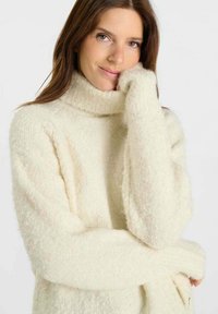 Jonge vrouw met lang bruin haar, gekleed in een crèmekleurige, zachte turtleneck-trui, die haar gezicht op haar hand steunt en zachtjes glimlacht.