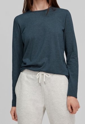 Frau in einem dunkelblauen Langarmshirt und hellgrauen Sweatpants mit einem weißen Kordelzug, stehend vor einem grauen Hintergrund.