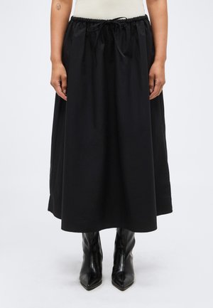 Personne portant une jupe noire longueur cheville avec une taille à cordon et des bottes à talons noires à bout pointu, debout devant un fond uni.