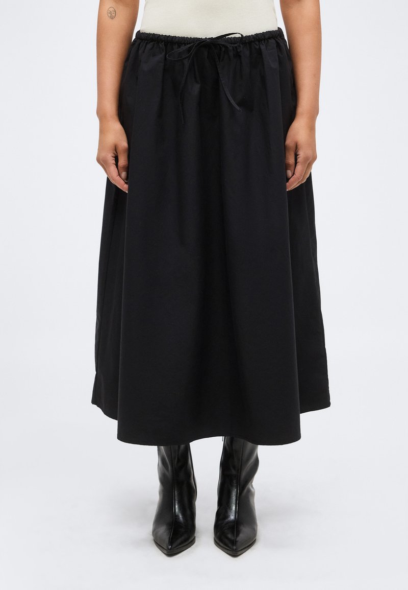 Άτομο που φοράει μαύρη φούστα μέχρι τον αστράγαλο με κορδόνι στη μέση και μαύρα ψηλοτάκουνα μποτάκια με μυτερή μύτη, στέκεται μπροστά σε απλό φόντο.