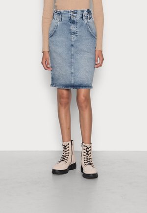 Personne portant une jupe en denim bleu délavé jusqu'aux genoux, un haut à manches longues côtelé beige et des bottines beige à lacets avec des semelles noires.