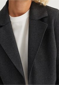 Blazer grigio scuro con rever a punta, realizzato in un tessuto texturizzato. Indossato sopra una t-shirt bianca a girocollo. Dettaglio sul colletto visibile.