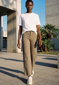Osoba chodící venku, která má na sobě bílé tričko, béžové široké kalhoty a bílé tenisky, s betonovými konstrukcemi a palmami v pozadí.