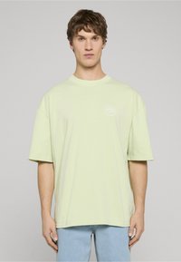 Světle zelené prodloužené tričko z bavlny s krátkými rukávy a kulatým výstřihem. Na hrudi je viditelné malé bílé logo.