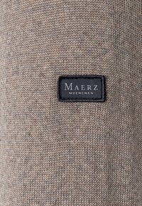 Strickpullover in Hellbraun mit glatter Textur, versehen mit einem schwarzen Stoffetikett, auf dem "Maerz Muenchen" steht.