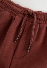 Lähikuva viininpunaisista collegehousujen vyötäröstä, jossa on elastinen kokoaminen ja sidottu punottu nyöri muovipäillä.