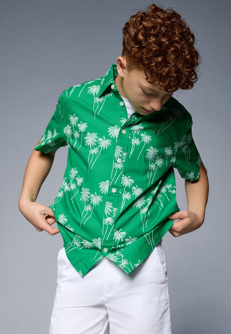 Camicia verde a maniche corte con stampa di palme bianche, chiusura a bottoni e orlo curvo. Indossata sopra una maglietta bianca e abbinata a pantaloni corti bianchi.