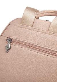 Zbliżenie różowego plecaka o fakturowanej powierzchni z beżowym paskiem i metalowym suwakiem na zewnętrznej kieszeni zapinanej na zamek.