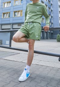 Grön Nike-tröja och matchande shorts med en logotyp. Vita och blå höga sneakers. Urban miljö med betong och byggnader.