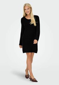 Robe noire en tricot avec des manches longues, un col rond et un ourlet côtelé. Modèle portant des talons bordeaux au design structuré et à bout pointu.