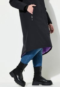 Svart lång rock med knappstängning, sidofickor och lila foder. Parat med blå jeans och svarta kängor med tjock sula.