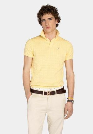 Giovane uomo che indossa una polo gialla a maniche corte, pantaloni beige, cintura scura, orologio e braccialetti, in piedi con una mano in tasca davanti a uno sfondo bianco.