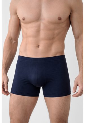 Torace maschile muscoloso che indossa boxer aderenti blu scuro in piedi contro uno sfondo chiaro e uniforme.