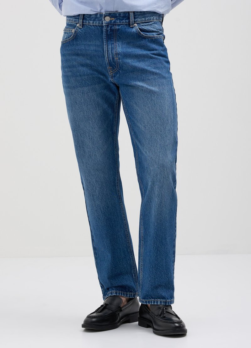 Jean en denim bleu avec une coupe droite, cinq poches et des détails de couture visibles, associés à des mocassins noirs.